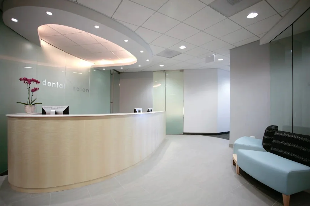 Reception area at Dental Salon: a curved front desk in light wood with a white countertop, the Dental Salon name on a frosted glass wall behind it, and a blue bench in the waiting area.